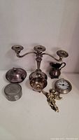 Full lot of seven vintage silver and metal items displayed on a white surface: three-arm candleholder, pitcher, covered dish, clock, small container, cherub figurine, small bowl.