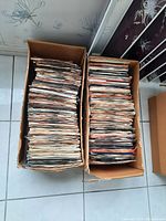 Two full boxes of 45 RPM vinyl records viewed from above on tiled floor