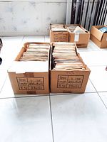Two cardboard boxes filled with 45 RPM vinyl records in paper sleeves, stacked on tile floor.