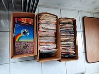 3 cardboard boxes filled with 45 RPM vinyl records, some record sleeves visible with colorful cover art and titles.