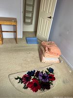 Image showing the demi-lune floral hooked rug on the carpet and the stacked blankets along the wall, plus a blue ortho cushion near the open door.
