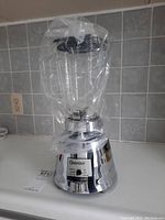 Osterizer blender with glass pitcher, plastic wrap on pitcher showing reflective chrome base with control buttons.