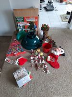 Photo showing the green swivel straight Christmas tree stand in front of its box, multiple colorful ornaments arranged in two rows, a red candle holder, candles, Christmas mini stockings including a Mickey Mouse plush stocking, pine and berry garland, and gift bags.