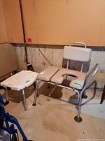 Photo of commode chair and shower stool side by side in basement with concrete floor and unfinished walls.