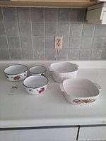 Two white ceramic CorningWare casserole dishes with fruit patterns and three white enamel metal food storage bowls with various fruit designs, displayed on a white countertop against a gray tiled backsplash.