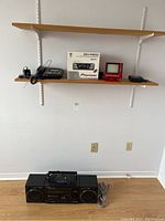 Wide view of shelf with electronics including boxed Pioneer DEH-P3500 car stereo, Cybiolink phone, small red CRT TV and other devices, with Panasonic stereo on floor below