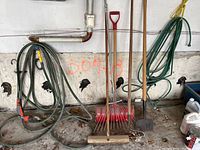 Garden tools including broom, two rakes, shovel, hoe, and hanging green garden hoses against a concrete wall.