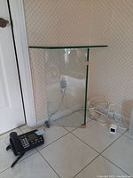 Glass hallway table positioned in corner near door, showing side view and bottom structure with visible cords around base.