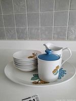 Eight small bowls stacked on large plate with sugar bowl with blue lid and creamer on top displaying the floral pattern of the set.