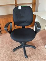 Front view of the black ergonomic office chair showing padded seat, backrest, and armrests on a carpeted floor with wooden furniture in the background.
