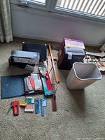 Photo showing garbage bin, shredder, rulers, stapler and other items set on carpet near window