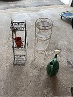 Photo showing the black metal plant stand with terracotta pot, white painted metal plant stand with three wicker-style round tiers, and green plastic watering can on a concrete floor.