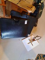 Two black portfolio cases and one metal armature with wooden base on floor near brown wooden easel.
