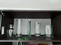 Five clear glass vases of various shapes and heights arranged on a dark shelf.