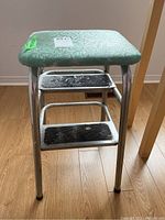 Front angle view of vintage metal step stool showing vinyl seat and two metal steps with wear.