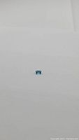 A small emerald cut blue gemstone on a plain light background, showing the stone's rectangular shape and blue color.