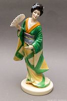 Front view of standing geisha figurine in green and yellow kimono holding a fan on circular base.