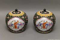 Front view of the two Asian hand painted ginger jars showing detailed figure scenes and colorful floral patterns on a black background.