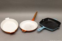 Three cast iron enamel cookware pans: oval, round with wooden handle, and square grill pan, all showing some wear.