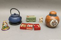 All items arranged in one photo showing the cast iron teapot with trivet, enamel bell, ginger jar, three boxed masks, green box, and carved figure.
