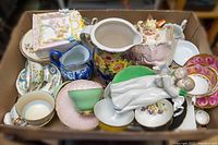 Box filled with assorted chinaware including teacups, saucers, various pitchers, and ceramic figurines all visibly used and some showing wear.