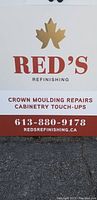 Photo of the Red's Refinishing advertising board with company logo, contact number, website, and details about cabinetry and crown molding repair services.