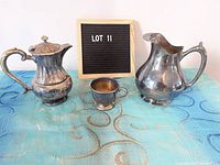 Three silverplate serving items including a coffee pot, creamer, and small cup shown on blue patterned cloth with lot sign.
