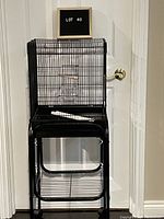 Front view of black metal bird cage on stand with rollers, showing cage dimensions and door with feeding cup and perch inside.