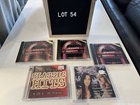 Five music CD cases displayed on a table with a lot 54 sign. Includes three 'Happy Days Rock and Roll' CDs, one 'All-Time Classic Hits 2-CD set,' and one 'Women & Song' compilation CD.
