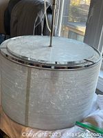 Cylindrical drum shade metal ceiling mount light fixture, showing top and side views, mounted on cloth surface by window. Shade fabric is translucent with bubble design.
