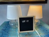 Two transparent glass table lamps with mushroom shaped bases and off-white drum shades shown on a blue patterned cloth. One lamp is turned on.