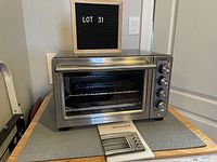 Front view of stainless steel KitchenAid toaster oven with glass door, four control dials, wire rack inside, and original user manual placed beneath it.