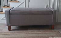 Front view of gray upholstered storage ottoman bench showing tufted cushion top, hinged lid, and wooden legs.