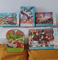 Five puzzle boxes arranged on a surface. Top row shows Molokini Current Sea Turtles, Dragon Flight, Realm of the Unicorn 350-piece Cobble Hill puzzles, and bottom row shows Crocodile Creek Land of Dinosaurs children's floor puzzle and Cobble Hill Snowmen Carollers puzzle 350-piece box.