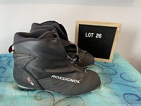 Side view of pair of black Rossignol X3 cross country ski boots on blue patterned surface with lot number board behind