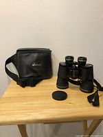 Full view of binoculars and original carrying case on wooden surface showing overall condition and presence of lens cover for one side missing.