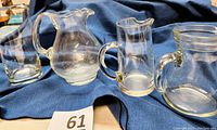 Four vintage glass pitchers on blue cloth, showcasing variety of shapes and handles, clear and etched frost glass included.
