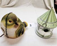 Photo showing side views of the bird-shaped birdhouse and the green and white cottage birdhouse with their hanging chains and perches arranged on a white backdrop.
