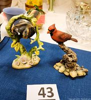Two ceramic bird figurines on a blue cloth. Left is a Flicker bird on a green branch base with pink flowers. Right is a red cardinal bird on a rock and branch base.