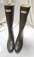 Front angled view of both tall olive green Hunter Wellington boots, showing iconic red and white Hunter logo on front top of boots.