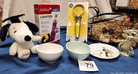 Photo showing Sunbeam nail trimmer in original box, JW shedding blade in packaging, plush Snoopy, stoneware pet bowls, cat-shaped earring holder, tapestry purse, Norman Rockwell plate, and cat figurine all displayed together.