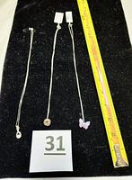 Three silver necklaces laid out on black fabric with a measuring tape showing about 18 inches length, displaying butterfly, infinity knot, and twisted infinity pendants.