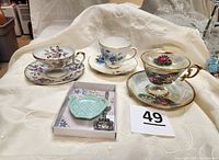 Three footed tea cup and saucer sets with floral designs and gold accents displayed on white fabric.