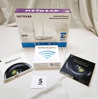 Four unopened boxed items: Netgear N300 WiFi Router box with two antennas pictured, Apple Aperture 2 and Aperture 3 software boxes, and Apple AirPort Express box with Wi-Fi icon.