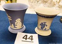 Two Wedgwood Jasperware urns on a blue cloth background: blue with white cherub neoclassical design and white with blue floral urn design.
