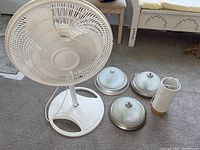 White oscillating fan with round base and three ceiling light fixtures with frosted glass covers and metallic rims alongside a tall white lamp with UK outlet plug.