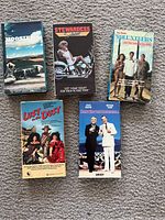Photo of five VHS tapes laid out on carpet showing front covers of Hoosiers, Stewardess School, Volunteers, Lust in the Dust, and Dirty Rotten Scoundrels