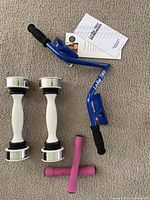 Blue Lo-Bak Trax spinal traction device with black handles, two white and silver shakable barbells, two small pink hand weights on carpeted floor with papers behind