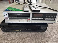 Front full view of Mitsubishi Betacord Beta player on top of black Sanyo VHS player with two remotes on top.