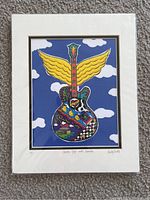 Photo of the full framed art print, showing a colorful guitar with wings against blue sky and clouds.